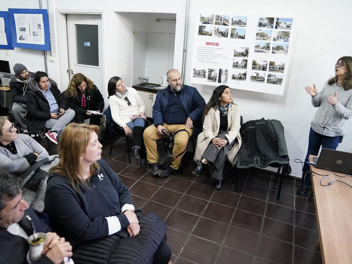 Reunión de vecinos y vecinas en un salón comunal con una moderadora dirigiendo la asamblea
